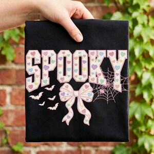 以下が含まれることがあります： 黒いTシャツに、パステルカラーの小さなハートで埋め尽くされた大きな白い文字で「SPOOKY」と書かれています。文字の下には、パステルカラーのハートが描かれた大きな白いリボン、3匹の小さな白いコウモリ、そしてクモの巣と小さなクモがデザインされています。