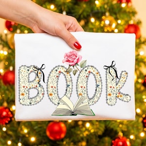 May include: A white t-shirt featuring the word BOOK in large letters filled with a floral pattern, outlined in black lace trim with small black bows. A pink rose sits above the letters, and an open book is illustrated at the bottom center.