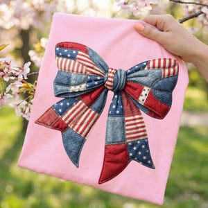 Puede incluir: Una camiseta rosa claro con un gráfico de un lazo grande en el centro. El lazo está construido con un diseño de patchwork de segmentos de tela roja, blanca y azul, alternando entre secciones azules con estrellas y secciones a rayas rojas y blancas.
