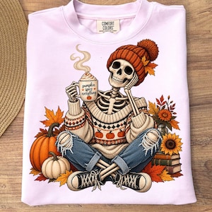Può includere: Una felpa rosa chiaro con la grafica di uno scheletro che indossa un berretto arancione e un maglione a motivi, che tiene in mano una tazza con la scritta pumpkin spice everything. Lo scheletro è seduto tra zucche, girasoli e foglie autunnali.