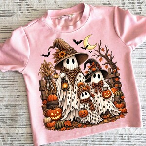 May include: A light pink t-shirt featuring a graphic print of three ghosts wearing witch hats and scarves. The ghosts are surrounded by pumpkins, a wooden fence, autumn leaves, bats, and a crescent moon. The design includes a lantern.