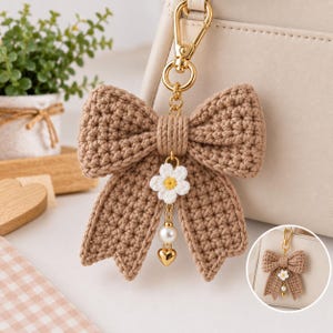 May include: A tan crocheted bow keychain featuring a gold-tone metal lobster clasp. A small white crocheted flower with a yellow center hangs from the center of the bow, followed by a white pearl bead and a small gold-tone metal heart charm.