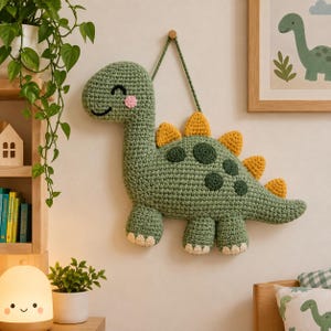 Può includere: Una decorazione da parete a forma di dinosauro lavorato all'uncinetto in verde, con collo lungo, un volto sorridente con guance rosa e una fila di punte triangolari gialle sul dorso. Il dinosauro presenta macchie circolari verde scuro e artigli beige.