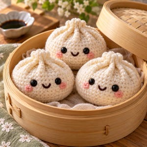 Peut inclure: Trois boulettes crochetées de couleur crème avec des yeux en perles noires, des sourires brodés et des joues cousues en rose, disposées dans un panier vapeur en bambou avec un couvercle assorti sur le côté.