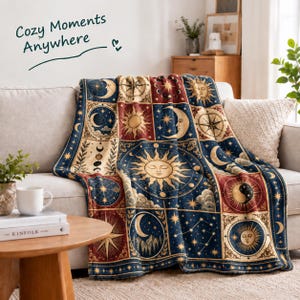 May include: A throw blanket featuring a grid of celestial illustrations including suns, moons, stars, and constellations in shades of navy blue, deep red, and gold. The text "Cozy Moments Anywhere" is printed in dark green above the blanket.