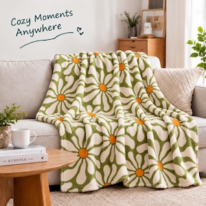 May include: A soft throw blanket featuring a repeating pattern of large white daisy flowers with bright orange centers on a sage green background. The text Cozy Moments Anywhere is printed in dark green script above the blanket.
