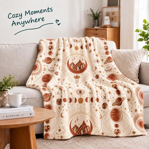 Peut inclure: Un plaid couleur crème orné d'un motif répétitif de symboles célestes orange rouille, incluant des planètes, des lunes, des soleils et des sommets montagneux dans des croissants. Le texte "Cozy Moments Anywhere" est imprimé en vert foncé.