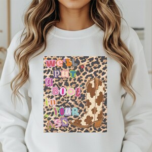 Puede incluir: Una sudadera blanca de cuello redondo con un estampado gráfico central sobre un fondo de leopardo. El diseño incluye una bota vaquera con estampado de vaca en marrón y blanco y un texto colorido con varios patrones que dice: "we'll put a boot in your ass."