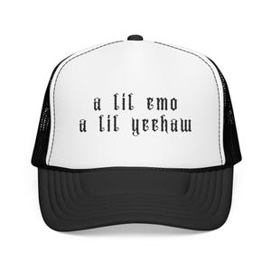 May include: A black and white trucker hat featuring a white front panel with the black text 'a lil emo a lil yeehaw' in a gothic font. The cap includes a black brim and black mesh side and back panels with a button on the top center.