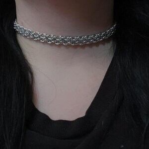 May include: A silver-toned chainmail choker necklace featuring a dense, woven metal link pattern. The jewelry piece sits closely around the neck, displaying a uniform, interlocking circular design with a polished, reflective metallic finish.