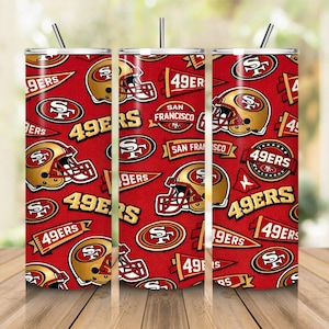 Puede incluir: Un vaso cilíndrico con pajita que presenta un patrón repetitivo de cascos de fútbol americano, banderines y logotipos de los San Francisco 49ers en rojo, dorado y blanco. El diseño incluye el texto 49ERS y SAN FRANCISCO impreso.