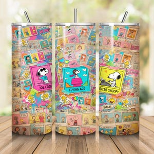 Peut inclure: Trois gobelets cylindriques avec couvercles et pailles, présentant un collage de bandes dessinées Peanuts. Les motifs incluent Snoopy en Joe Cool, Flying Ace et Writer Snoopy, avec des accents colorés, des autocollants et du texte sur un fond papier.