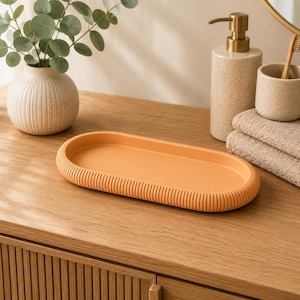 May include: An oval-shaped tray in a solid matte orange colour featuring a textured, ribbed edge design. The tray has a smooth, flat interior surface and a rounded, raised rim with vertical grooves running along the entire outer perimeter.