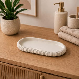 May include: An oval-shaped tray with a smooth, flat interior and a textured, ribbed exterior edge. The tray is off-white in colour and features a minimalist, clean design suitable for holding small items on a flat surface in a bathroom or living space.