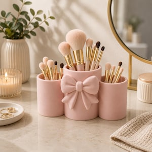 May include: A light pink makeup brush holder featuring three cylindrical compartments and a prominent bow detail on the centre section. The holder is filled with various makeup brushes that have gold-toned ferrules and soft, light-coloured bristles.