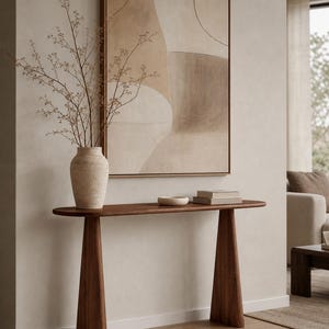 May include: A dark wood console table with a long oval top and two wide, tapered legs. On the table sits a textured beige vase containing thin, branching stems, a small shallow bowl, and two stacked books beneath a large framed abstract art piece.