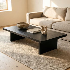 Puede incluir: Una mesa de centro rectangular de madera negra con superficie lisa. Sobre la mesa hay un cuenco de cerámica texturizada, un libro cerrado de tapa clara y una escultura de piedra beige abstracta con una abertura central.