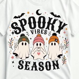 May include: A white t-shirt featuring a graphic print with the text SPOOKY VIBES SEASON. The design includes three ghosts wearing beanies in olive green, pink, and orange, surrounded by bats, stars, and small floral sprigs in black and orange.