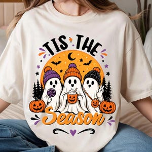 Puede incluir: Una camiseta de color crema con un gráfico de tres fantasmas con gorros coloridos. Los fantasmas están frente a una gran luna naranja con siluetas de árboles, murciélagos y estrellas. El texto dice "TIS THE Season" en negro y naranja.