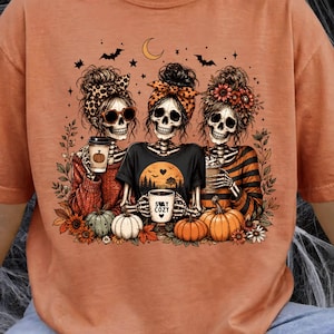 May include: A burnt orange t-shirt featuring a graphic of three skeletons with messy buns, wearing headbands and holding coffee cups. The central skeleton wears a shirt with a sunset design and the text STAY COZY. Pumpkins, leaves, and bats surround them.
