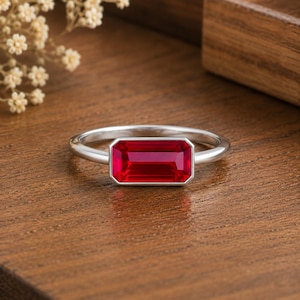 May include: A silver-toned ring featuring a single, rectangular, emerald-cut vibrant red gemstone set horizontally in a smooth bezel setting. The band is thin, polished, and circular, providing a clean and simple frame for the prominent red stone.