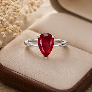 May include: A silver ring featuring a single pear-shaped, deep red gemstone set in a prong setting. The band is smooth and polished, showcasing a clean metal finish that complements the vibrant, faceted red stone centered on the ring band.