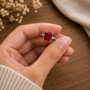 May include: A silver-toned ring featuring a large, rectangular, emerald-cut red gemstone set in a four-prong setting. The vibrant red stone is positioned centrally on a smooth, polished metal band, showcasing a clear, faceted surface design.