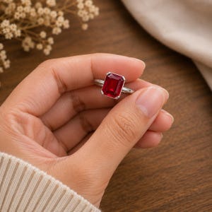 May include: A silver-toned ring featuring a large, rectangular, emerald-cut red gemstone set in a four-prong setting. The vibrant red stone is positioned centrally on a smooth, polished metal band, showcasing a clear, faceted surface design.