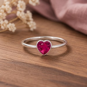 Könnte beinhalten: Ein silberner Ring mit einem einzelnen herzförmigen, leuchtend pinkfarbenen Edelstein, der in einer glatten Metallfassung sitzt. Das Band ist dünn und poliert und zeigt ein klares Design, das den zentralen herzförmigen Stein hervorhebt.