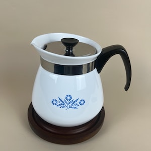 May include: A white teapot with a blue floral pattern on the front, featuring a black handle, a stainless steel lid with a black knob, and a silver metal band around the top. The teapot is resting on a circular dark brown wooden base.