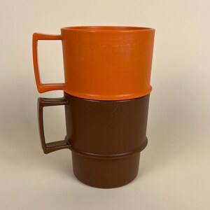 May include: Two stackable mugs with square-shaped handles. The top mug is a solid bright orange color, while the bottom mug is a solid dark brown color. Both mugs feature a cylindrical body with a distinct horizontal ridge around the middle section.
