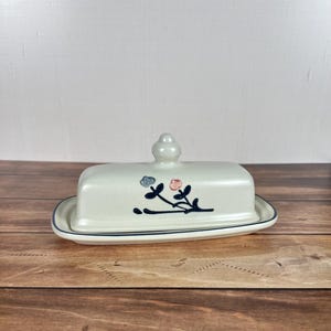 May include: A cream-colored ceramic butter dish with a rectangular base and a matching lid featuring a central knob. The lid is decorated with a hand-painted dark blue floral vine motif, accented by a single light blue flower and a soft pink flower.