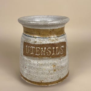 May include: A cylindrical ceramic utensil crock with a speckled off-white glaze and brown accents. The word UTENSILS is embossed in white capital letters on a rectangular brown panel centered on the front of the vessel, which has a flared rim.