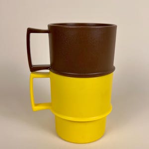 May include: Two stackable mugs with square handles, one brown and one yellow. The brown mug sits on top of the yellow mug, which features a stepped design that allows the upper mug to nest securely within the base of the lower mug. Both mugs have a smooth, solid-colored finish and a consistent, functional shape.