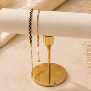 May include: A gold-toned metal jewellery display stand with a cylindrical, cream-coloured velvet bar. Two bracelets hang from the bar: one featuring a black and gold twisted cord design and the other a delicate gold-toned chain with a small clasp.