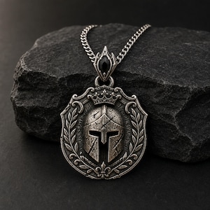 May include: A silver-toned pendant necklace featuring a circular medallion with a central Spartan helmet, a crown, and laurel leaf engravings. The pendant is suspended from a silver chain and includes a small, black, marquise-cut gemstone.
