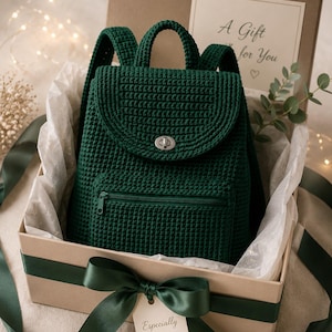 Puede incluir: Una mochila de ganchillo verde oscuro con asa superior, correas para los hombros, un bolsillo frontal con cremallera y un cierre giratorio de metal plateado. La mochila se presenta en una caja de regalo color canela con un lazo verde y una etiqueta Especially for you.