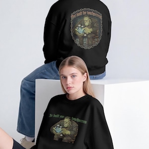 Puede incluir: Una sudadera negra de manga larga con un gráfico de un monstruo verde sosteniendo un objeto azul brillante. El texto "Not built for touchscreen" está impreso sobre el monstruo en un marco circular en la espalda y en línea recta en la parte delantera.