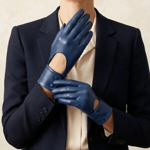 May include: A pair of navy blue leather gloves featuring a cutout detail on the back of the hand and a wrist strap secured with two matching buttons. The gloves are worn with a dark navy blue blazer and a light-colored button-down shirt underneath.