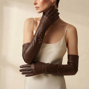 May include: A pair of long, dark brown leather gloves extending past the elbows. Each glove features a row of four matching dark brown buttons along the outer wrist area, providing a decorative detail on the smooth, fitted leather surface.