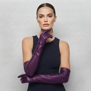 Könnte beinhalten: Ein Paar lange, tiefviolette Lederhandschuhe, die über die Ellbogen reichen. Die Handschuhe verfügen über eine Reihe kleiner, passender violetter Knöpfe entlang des äußeren Unterarms, was den Händen und Armen ein strukturiertes Aussehen verleiht.