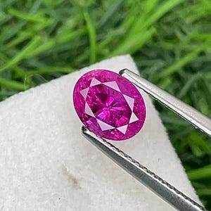 Könnte beinhalten: Ein leuchtender, oval geschliffener magentafarbener Edelstein, der von einer silberfarbenen Metallpinzette gehalten wird. Der Stein hat eine facettierte Oberfläche, die das Licht reflektiert und einen tiefen, gesättigten pink-violetten Farbton zeigt.