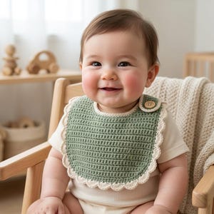 Könnte beinhalten: Ein salbeigrünes gehäkeltes Baby-Lätzchen mit cremefarbenem Muschelrand und einem einzelnen Holzknopf als Verschluss an der Seite. Das Lätzchen wird über einem schlichten, cremefarbenen Kurzarm-Body getragen und zeigt ein strukturiertes Maschenbild.