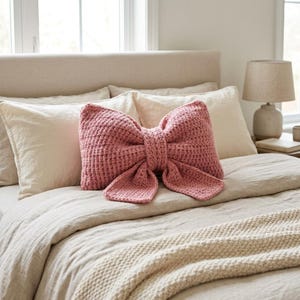 Op de afbeelding: Een oudroze decoratief sierkussen in de vorm van een grote strik. Het kussen heeft een textuur van gehaakt breiwerk met een centrale knoop en twee lange, rechthoekige uiteinden die vanuit het midden van de strik naar beneden hangen.