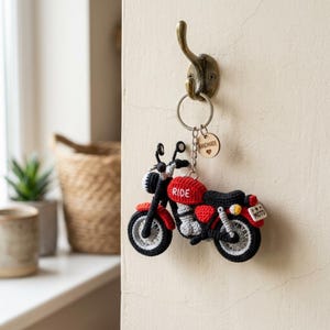 Może przedstawiać: Szydełkowany brelok do kluczy w kształcie motocykla z czerwonym zbiornikiem paliwa z białym napisem RIDE, czarnym siedzeniem, czarnymi kołami ze srebrnymi szprychami oraz małą białą tablicą rejestracyjną z napisem MOTO. Drewniana zawieszka ma napis HANDMADE.