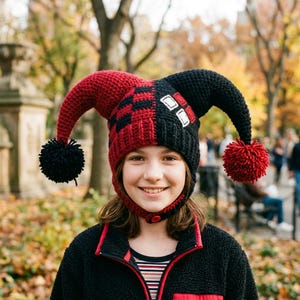 May include: A crocheted jester hat featuring two long, curved points with pom-poms at the ends. The hat is split into red and black sections with a checkered pattern on the red side and white square accents on the black side, secured by a button.