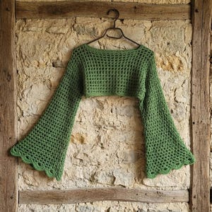 May include: A sage green long-sleeve crochet crop top featuring an open-knit mesh pattern, a wide boat neckline, and dramatic flared bell sleeves with a scalloped edge finish. The garment is displayed on a black wire hanger.