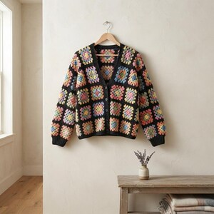May include: A long-sleeved cardigan featuring a black base with a colorful granny square pattern in shades of pink, yellow, blue, and green. The garment has a V-neckline, a front button closure, and ribbed cuffs and hem in solid black.
