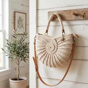 Pode incluir: Uma bolsa de crochê na cor creme em formato de concha com um centro em espiral. Possui uma alça de corda trançada grossa, uma alça de ombro longa e ajustável de couro bege e um pequeno pingente de estrela-do-mar em metal.