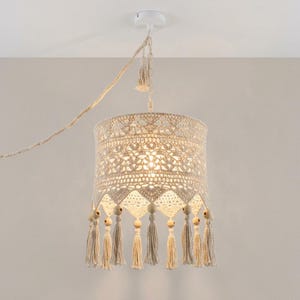May include: A cream-colored crochet pendant light fixture featuring an intricate lace pattern, a cylindrical shade, and a series of hanging tassels accented with round wooden beads. The lamp is suspended by a matching braided cord and chain.